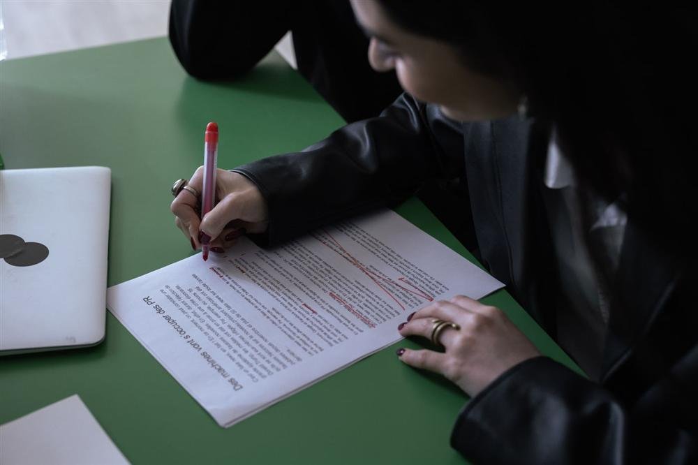Person using a red pen to edit a printed document next to a laptop